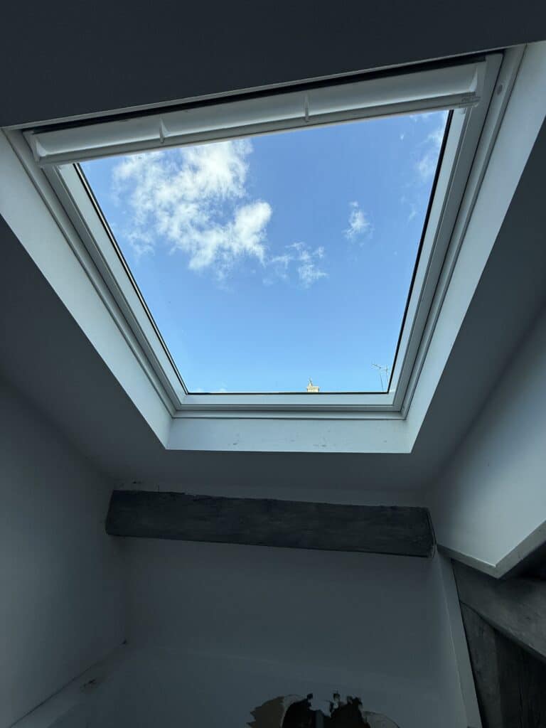 Fenêtre de toit Velux, ciel bleu nuageux et poutre en bois rustique.