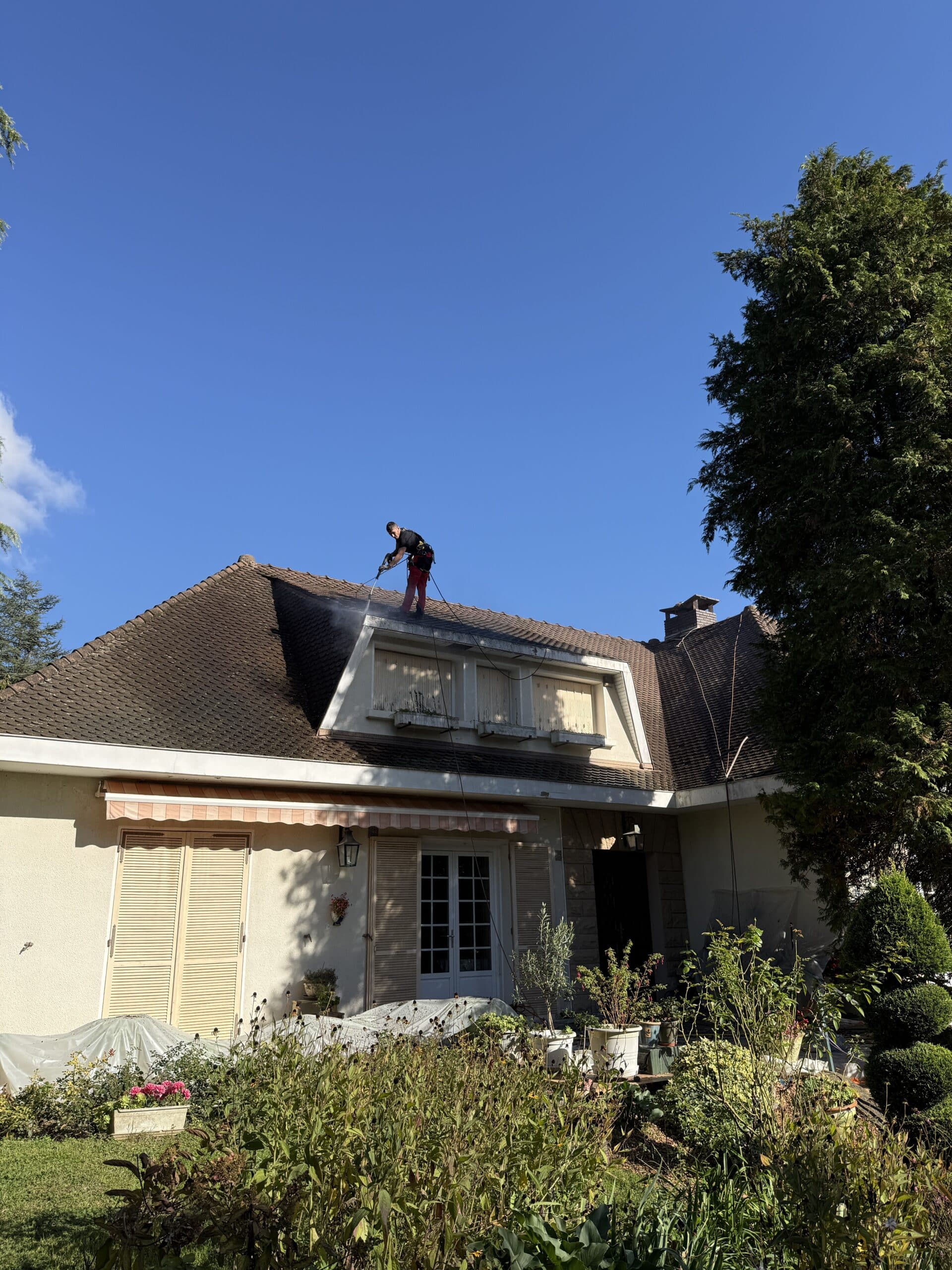 Nettoyage de toiture et démoussage professionnel de maison Artisan nettoyant au jet haute pression les tuiles d'un toit de maison sous un ciel bleu.